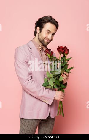 Ospite positivo di evento in giacca e cravatta di prua che tiene rose su sfondo rosa, immagine stock Foto Stock
