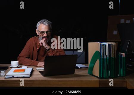 allegro uomo d'affari dai capelli grigi che guarda il laptop mentre ha una videochiamata di notte in ufficio, immagine stock Foto Stock