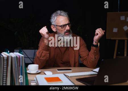 irritato uomo d'affari che tiene pugni clenched durante la videochiamata sul laptop in ufficio di notte, immagine di scorta Foto Stock