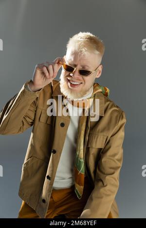 luce del sole sul volto di un uomo albino sorridente regolazione occhiali da sole eleganti su grigio, immagine stock Foto Stock