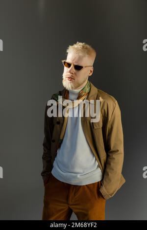 luce del sole sul volto di elegante albino uomo in occhiali da sole posa con le mani in tasche su grigio, immagine stock Foto Stock