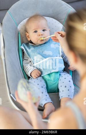 Il cucchiaio della madre che alimenta il bambino del suo bambino del bambino nella sedia del bambino con purea di frutta. Concetto di introduzione degli alimenti solidi per bambini Foto Stock