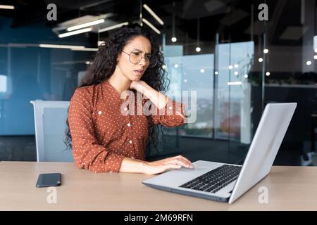 Donna d'affari troppo stanca che lavora in ufficio con un computer portatile, la lavoratrice ispanica ha un forte dolore al collo, massaggiando i muscoli con la mano. Foto Stock