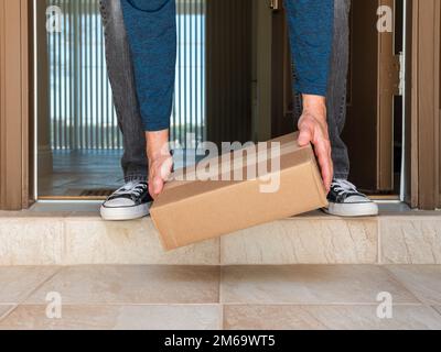 Uomo che raccoglie una scatola di imballaggio consegnata a una porta residenziale. Ordine online consegna pacchetto al portico anteriore di casa. Foto Stock