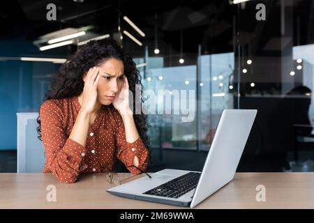 Donna ispanica troppo stanca che lavora all'interno di un ufficio moderno, lavoratore in ufficio con un computer portatile con mal di testa grave. Foto Stock