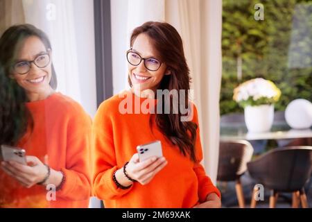 Attraente donna dai capelli lunghi che indossa un maglione arancione mentre si alza alla finestra e si guarda fuori. Messaggi di testo femminili allegri. Foto Stock