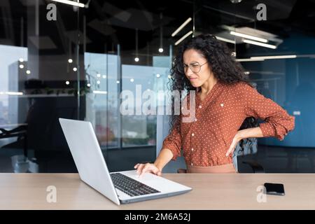 Donna ispanica stanca che lavora tardi, donna d'affari che soffre di mal di schiena grave, lavoratrice all'interno dell'ufficio seduto a un tavolo con un computer portatile al lavoro. Foto Stock