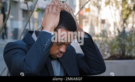 Stressato uomo etnico esausto ragazzo maschio azienda dipendente che ha dolore alla testa mal di testa stress mal di testa con perdita di lavoro licenziato disoccupazione in città. Stanco Foto Stock