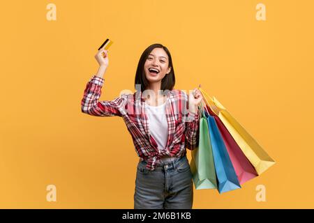 Shopping facile. Donna asiatica overjoyed che tiene la carta di credito e borse shopper luminoso, ottenuto bonus cashback, sfondo giallo Foto Stock