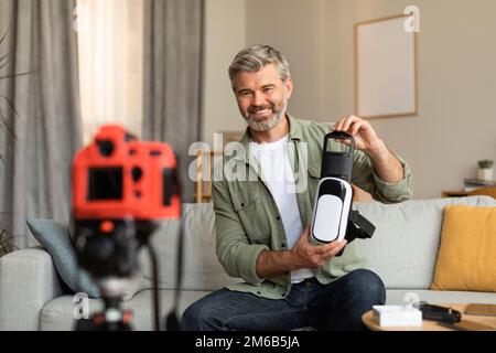 Sorridente anziano uomo caucasico ripresa video blog, mostrando occhiali vr per l'intrattenimento in salotto Foto Stock