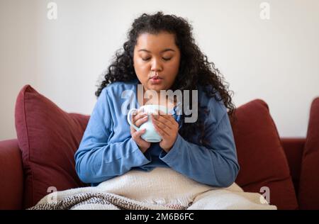 Donna coperta di coperta, seduta sul divano bere tè caldo Foto Stock