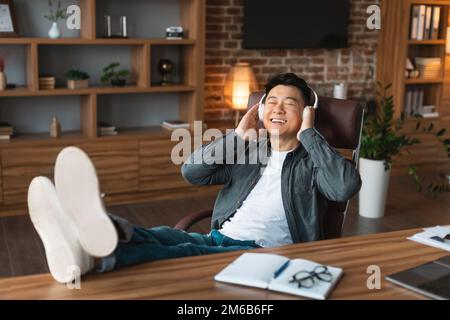 Ridendo maschio giapponese di mezza età in cuffie wireless gode la canzone, ascolta la musica sul posto di lavoro Foto Stock