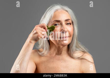 Femmina caucasica matura con capelli grigi fare massaggio di pietra di giada a casa, isolato su sfondo grigio, studio Foto Stock