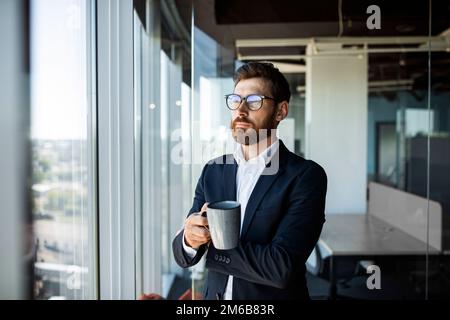 Uomo d'affari sognante che tiene la tazza del caffè, che guarda da parte e che gode il nuovo lavoro, in piedi vicino al posto di lavoro in ufficio Foto Stock