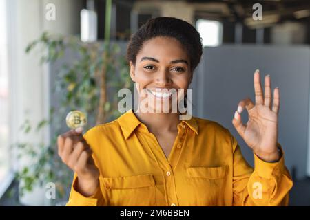 Una donna d'affari afroamericana di successo che tiene il bitcoin e che mostra un gesto ok, posando all'interno dell'ufficio, spazio libero Foto Stock