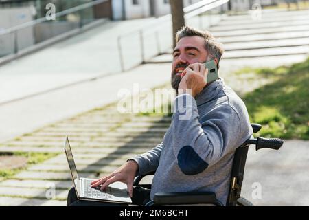 Uomo in sedia a rotelle che guarda lontano mentre parla al telefono e usa il computer portatile all'aperto. Foto Stock