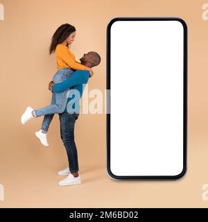 Amando sposi neri che posano vicino a grande smartphone vuoto con schermo bianco su sfondo pesca studio, mockup Foto Stock