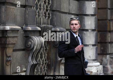 Bel giovane uomo in abito nero fumo sigaro piccolo sulla strada Foto Stock
