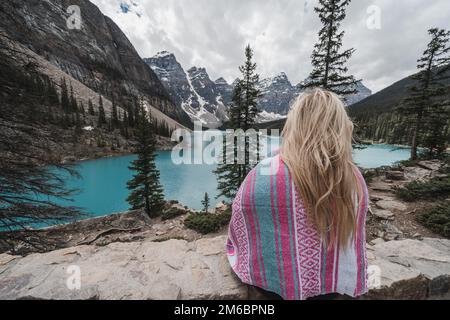 Vista posteriore di una donna bionda coccolata in una coperta rosa, guardando il lago Moranie in Canada Foto Stock