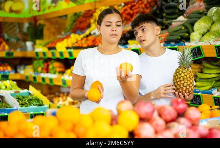 Giovane coppia positiva scegliendo ananas freschi e arance frutta tropicale in supermercato Foto Stock