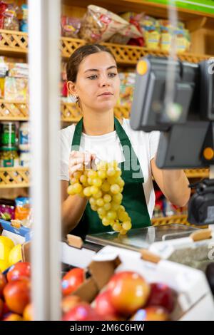 Giovane lavoratrice di supermercati che pesano sulle scale uve verdi in negozio di alimentari Foto Stock