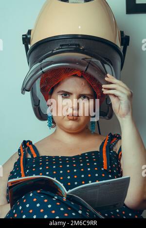 giovane donna d'epoca ispanica latina di etnia argentina, al parrucchiere facendo arricciacapelli su una macchina arricciacapelli, con una rivista in mano e lo Foto Stock