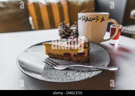 Dessert e una bevanda calda in una caffetteria. Un pezzo di torta decorata con zucchero a velo e frutta posta su un piatto nero. Una tazza con una bevanda calda sfocata sullo sfondo. Foto di alta qualità Foto Stock