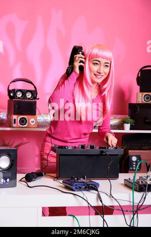 Asian dj mixare musica elettronica sui giradischi mentre si gioca record mix suoni in studio con sfondo rosa. Performer divertirsi mentre si esibisce una canzone techno, godendosi lo stile di vita notturno Foto Stock