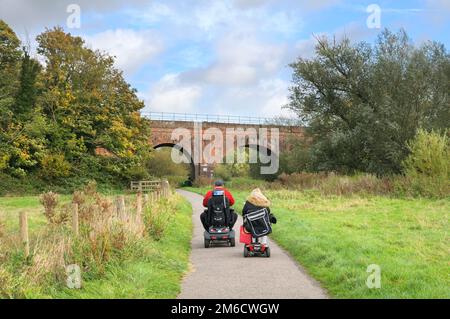 Anziani in sella a scooter per la mobilità su un percorso principale attraverso Hambrook Marshes, che si avvicina a un ponte ferroviario ad arco, Canterbury, Kent, Regno Unito Foto Stock