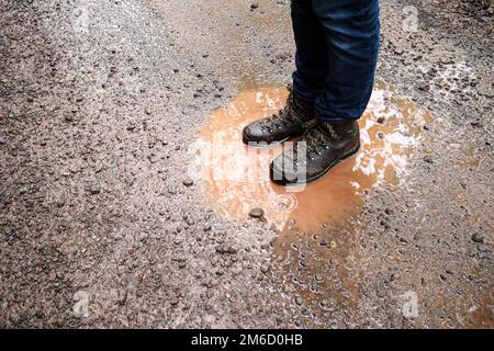 Gambe inferiori dell'uomo in jeans blu e piedi in scarpe da trekking, in piedi in pozza fangosa su strada sterrata dopo una pioggia. Aspetto grintoso e sgranato. Foto Stock