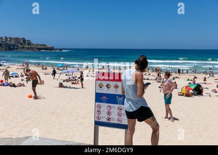 Bondi Beach Sydney 2023, giovane donna in brunetta che invia sms sul suo telefono cellulare, sui tatuaggi delle gambe e delle braccia, si trovava accanto al cartello di Bondi Beach, Sydney, NSW, Australia Foto Stock