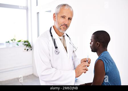 Fa sentire i suoi pazienti a suo agio. Ritratto di un medico di sesso maschile maturo in piedi con una siringa con il paziente sullo sfondo. Foto Stock