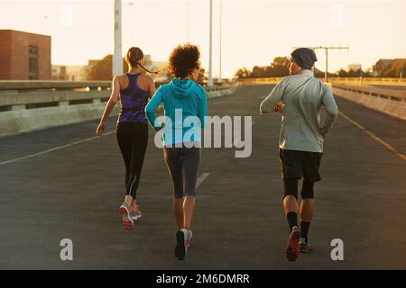 Correre in una città tranquilla. Ripresa da dietro di tre giovani joggers che corrono lungo una strada vuota all'alba. Foto Stock