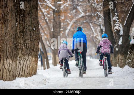 Padre e bambini in bicicletta in inverno. Un uomo con suo figlio e sua figlia cavalcano le biciclette lungo la pista ciclabile nel parco invernale. Vista dal retro Foto Stock