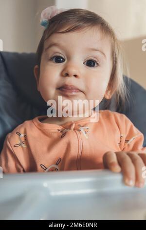 Primo piano ritratto di bambina seduta su una sedia alta pronto a mangiare, in attesa di cibo a pranzo. Famiglia felice. Bellezza viso. Foto Stock