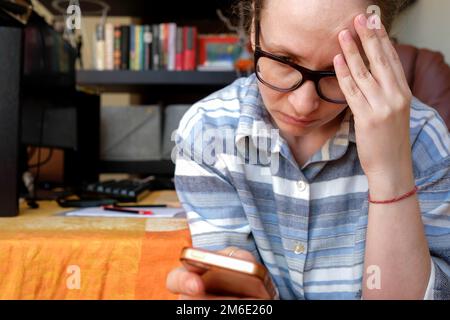 Legge le cattive notizie dal telefono. Una giovane donna sta tenendo la testa. Negativo online Foto Stock