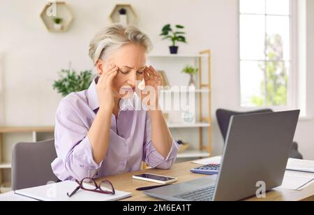 Donna con capelli grigi infelice soffre di mal di testa e emicranie si siede a un tavolo con un notebook in ufficio Foto Stock