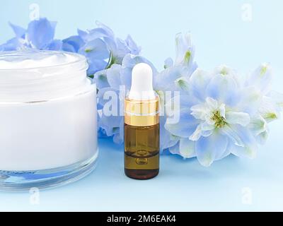 Siero in bottiglia di vetro oro con pipetta o olio essenziale, crema idratante in vaso di vetro aperto e fiore blu di Delphinium. Cosmetologia idratante bea Foto Stock