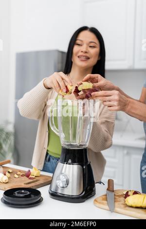 Donna che mette la mela nel frullatore vicino sfocato amico asiatico in cucina, immagine stock Foto Stock