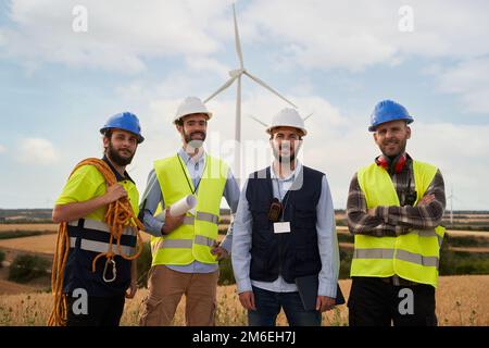Ritratto di un gruppo di ingegneri che guarda la telecamera in un campo agricolo con turbine eoliche Foto Stock