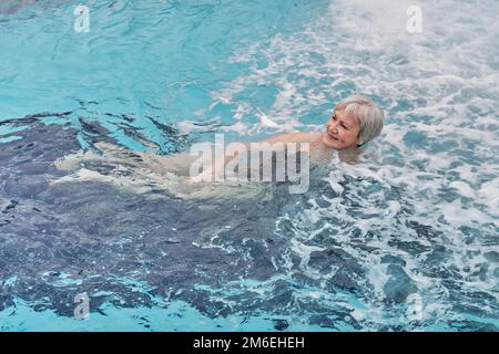 Donna anziana sorridente felice con capelli corti grigi guardando la macchina fotografica nella piscina termale esterna con idromassaggio. Concetto seniors attivo. Foto Stock