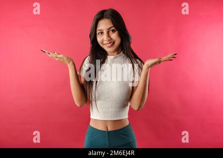 Giovane ragazza dai capelli scuri sorridente felice indossando il raccolto a coste isolato su sfondo rosso che ruggisce e getta le mani da parte con clueless. Non so cosa Foto Stock