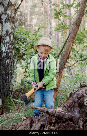 Bambino in un cappello nella foresta autunnale. Un ragazzo con un'ascia taglia un albero. Piccolo lumberjack. Lavori nel bosco. Abbattimento di alberi. Foto Stock