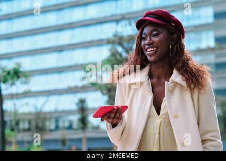 Donna elegante che guarda lontano e sorridente mentre tiene un telefono cellulare all'aperto. Foto Stock