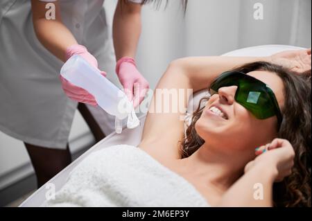 Foto ravvicinata delle mani di estetista che applicano il gel alla spatola prima di lavorare con la macchina per la rimozione dei peli laser. Bella cliente sorridente sdraiato con le braccia sollevate durante la sessione di capelli senza laser. Foto Stock