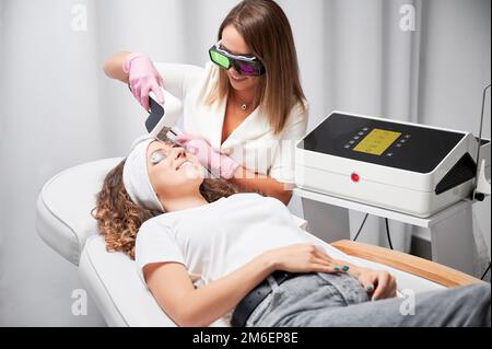 Donna sorridente in occhiali protettivi che riceve un trattamento laser del viso in salone di bellezza. Cosmetologa femminile che utilizza un dispositivo di scansione laser a diodi durante la procedura di rifacing skincare. Foto Stock