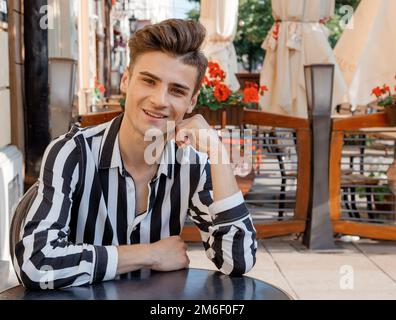 Modello ragazzo vicino al caffè. Un giovane uomo in abiti eleganti è seduto a un tavolo. Carino ragazzo sulla terrazza estiva con fiori. Foto Stock