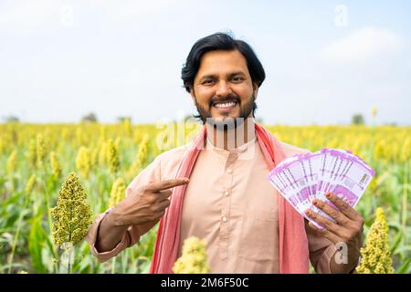 Felice giovane agricoltore sorridente mostrando denaro o valuta puntando il dito mentre si guarda la fotocamera a terreno agricolo - concetto di guadagno, profitto agricolo Foto Stock