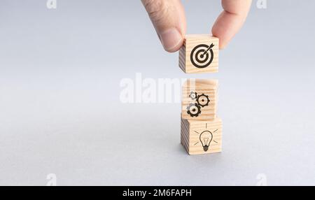 Strategia aziendale e concetto di piano d'azione. Mano mettere blocco di legno con l'icona bersaglio sopra gli altri con gli ingranaggi e le icone lampadina. Sfondo grigio. Foto Stock