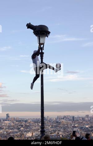 Street performer a Parigi Montmartre sale il lampione e colpisce una palla. Lo skyline della città di Parigi è sullo sfondo Foto Stock